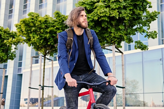 Bebaarde hipster man met rugzak zit op de rode vaste fiets in een park.