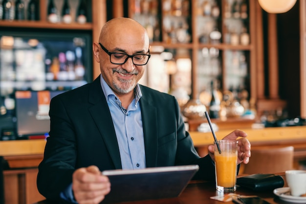 Bebaarde hogere volwassene in kostuum die sap drinken en tablet gebruiken terwijl het zitten in cafetaria.