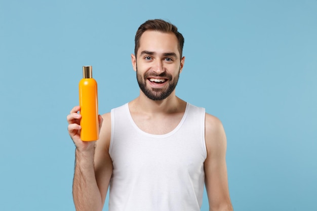 Bebaarde jonge man 20 jaar oud in wit overhemd houd fles shampoo geïsoleerd op blauwe pastel muur achtergrond, studio portret. Huidverzorging gezondheidszorg cosmetische ingrepen concept. Mock up kopie ruimte.