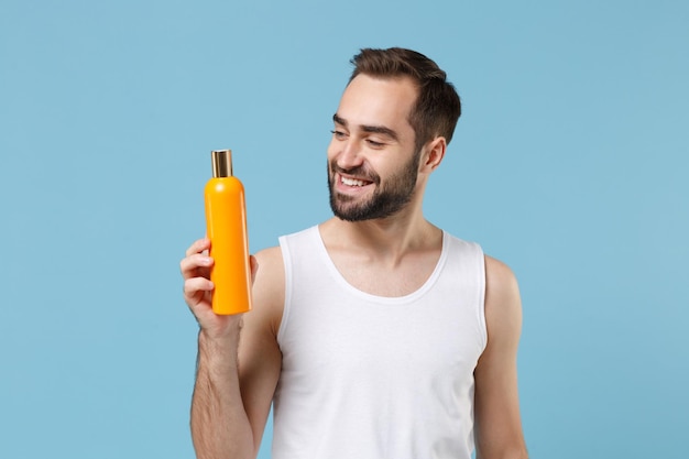 Bebaarde jonge man 20 jaar oud in wit overhemd houd fles shampoo geïsoleerd op blauwe pastel muur achtergrond, studio portret. Huidverzorging gezondheidszorg cosmetische ingrepen concept. Mock up kopie ruimte.