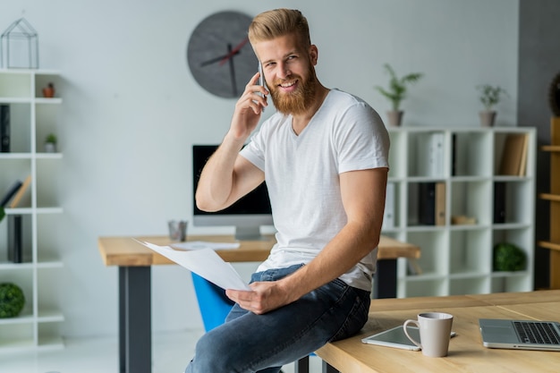 Bebaarde jonge zakenman werken bij moderne kantoor. man met wit t-shirt en het maken van aantekeningen op de documenten.