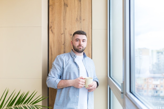 bebaarde knappe man staat bij raam met een kopje in zijn hand op het balkon en drinkt een warme koffie of thee man in vrijetijdskleding geniet van doorkijk op kantoor of thuis ontspannen of time-out op het werk