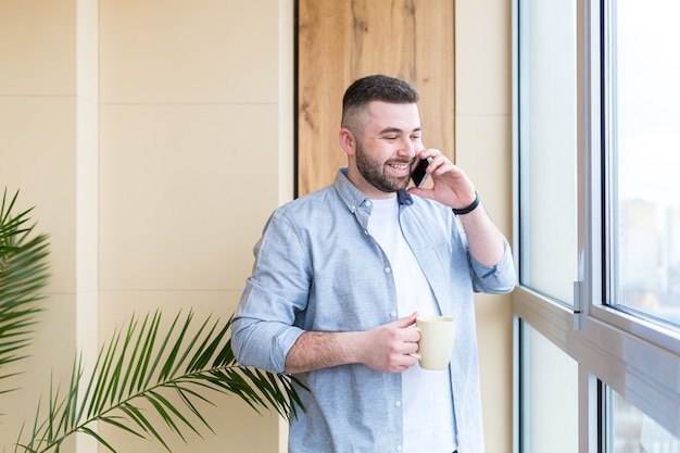 bebaarde knappe man staat bij raam met een kopje in zijn hand op het balkon en drinkt een warme koffie of thee man in vrijetijdskleding geniet van doorkijk op kantoor of thuis ontspannen of time-out op het werk