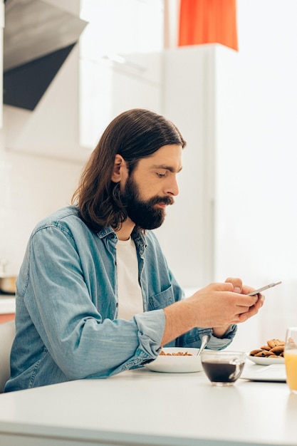 Bebaarde langharige blanke man zit in de keuken met een kom cornflakes en een kopje koffie en gebruikt een smartphone