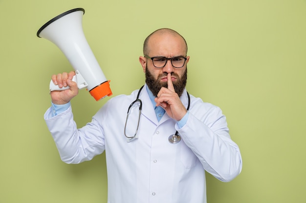 Bebaarde man arts in witte jas met een stethoscoop om de nek dragen van een bril met megafoon met ernstig gezicht stilte gebaar maken met vinger op lippen