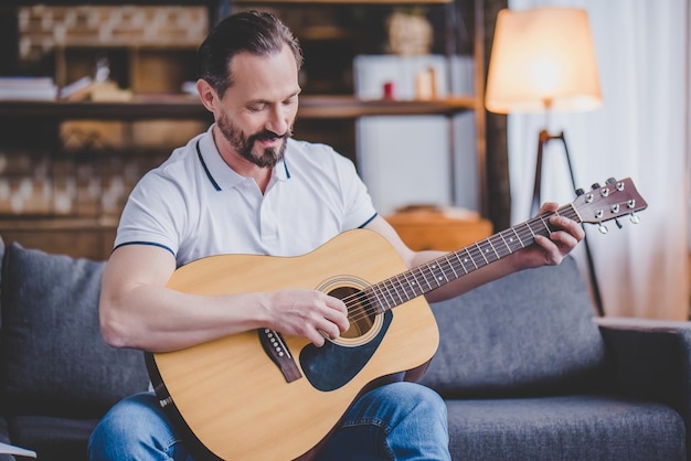 Bebaarde man die gitaar speelt terwijl hij thuis op de bank zit