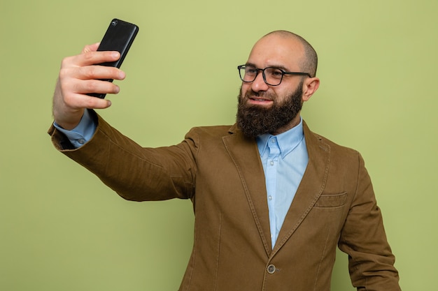 Bebaarde man in bruin pak met een bril die selfie maakt met een smartphone die vrolijk lacht over een groene achtergrond