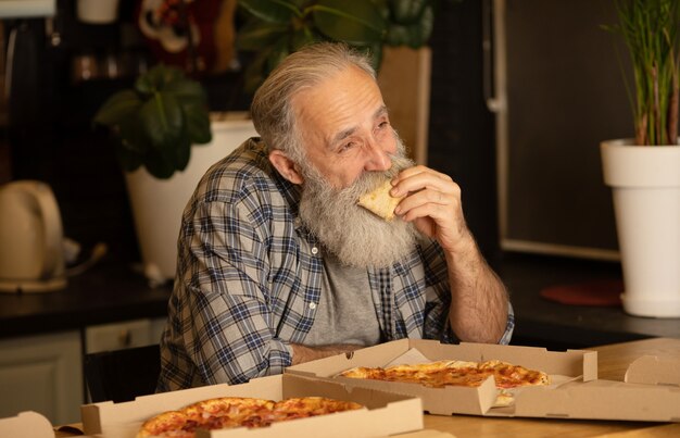 Bebaarde man in een keuken. Een senior man in een blauw shirt. Man thuis met pizza.