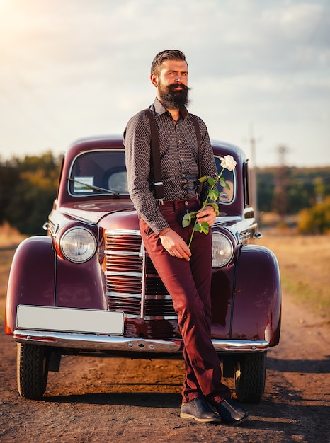 Bebaarde man in een shirt, broek met bretels houdt een witte roos in zijn hand in de buurt van een bruine retro auto op een landweg en kijkt op zijn horloge
