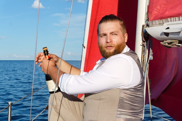 Foto bebaarde man in een vest met een fles mousserende wijn naast de mast op een zeilboot