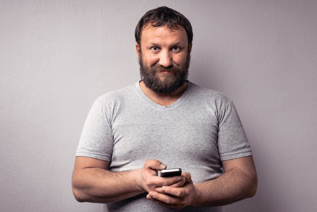 Bebaarde man in grijs t-shirt met mobiele telefoon terwijl hij tegen een grijze muur staat