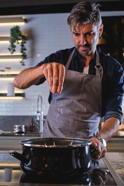 Bebaarde man in grijze schort die zout in de pan doet tijdens het bereiden van voedsel in de moderne keuken