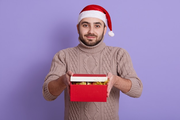 Bebaarde man in kerstmuts en beige trui brengt geschenken in rode doos, poseren geïsoleerd op paars