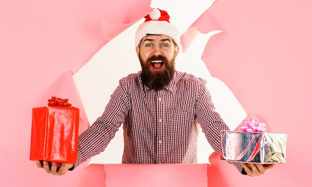 Bebaarde man in kerstmuts en geruit overhemd met cadeautjes, geschenkdozen die door een papieren gat kijken