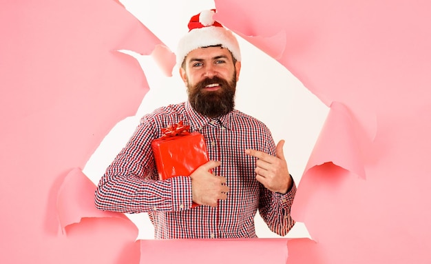 Bebaarde man in kerstmuts met huidige stijlvolle man die met vinger op seizoensverkoop van geschenkdoos laat zien