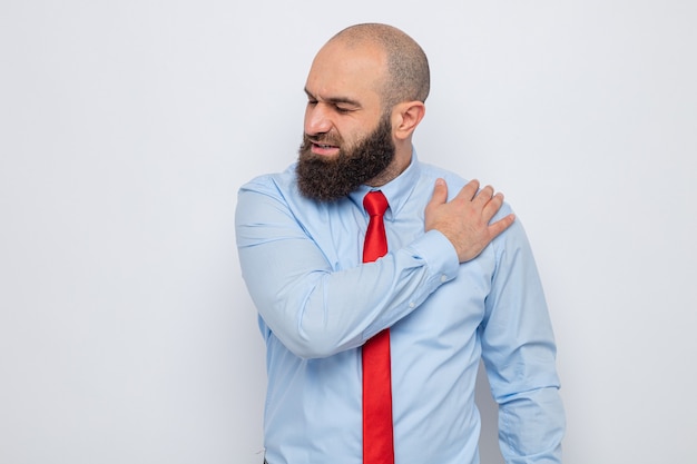 Bebaarde man in rode stropdas en blauw shirt die er onwel uitziet en zijn schouder aanraakt, voelt pijn terwijl hij op een witte achtergrond staat