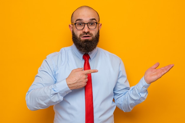 Bebaarde man in rode stropdas en blauw shirt met bril kijkend naar camera met zelfverzekerde glimlach presenteren met arm van haar hand wijzend met wijsvinger naar haar hand staande over oranje achtergrond