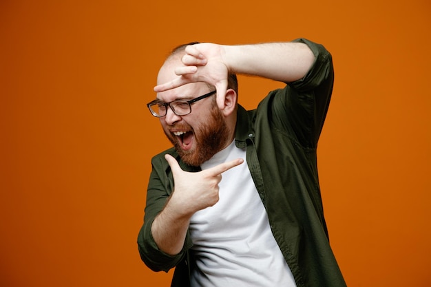 Bebaarde man in vrijetijdskleding met een bril die een frame maakt met vingers die door dit frame naar de camera kijken, blij en opgewonden over oranje achtergrond