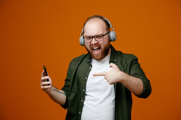 Bebaarde man in vrijetijdskleding met een bril met koptelefoon die een smartphone vasthoudt die met de wijsvinger erop wijst en er opgewonden en gelukkig uitziet over een oranje achtergrond