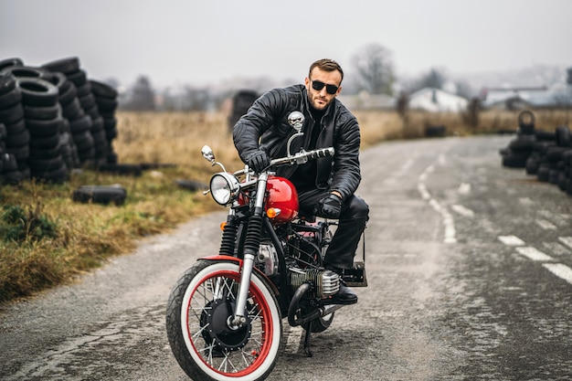 Bebaarde man in zonnebril en lederen jas kijken naar de camera zittend op een motorfiets op de weg. Achter hem staat een rij banden