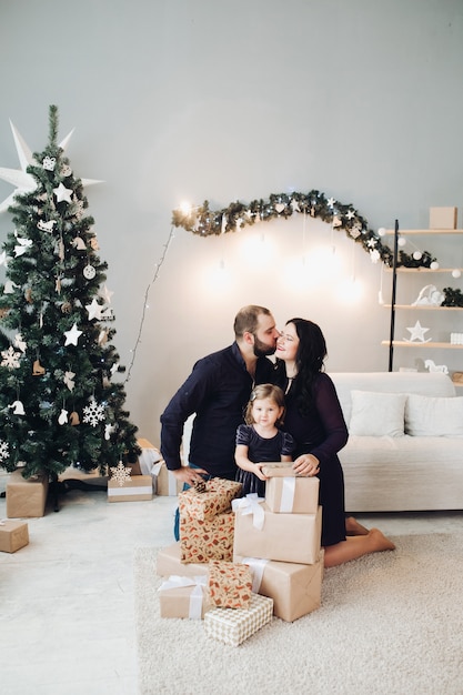 Bebaarde man kussende vrouw terwijl dochter het stellen. Familie dragen in zwarte kleding. Woonkamer versierd met kerstboom, dennenslinger, lichtsnoer, geschenkdozen.