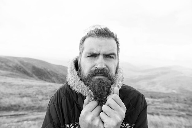 Bebaarde man lange baard brutale Kaukasische hipster met snor op ernstig gezicht ongeschoren man met stijlvol haar krijgen baarden kapsel ineenkrimpen van de kou op de bergtop op natuurlijke bewolkte hemelachtergrond