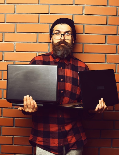 Bebaarde man lange baard brutale Kaukasische verrast ongeschoren hipster met laptops in rood zwart geruit overhemd met hoed en bril op bruine bakstenen muur studio achtergrond