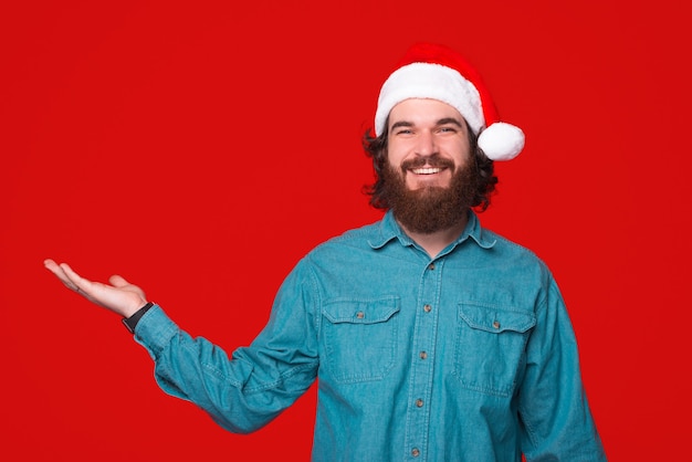 Bebaarde man met kerstmuts presenteert iets met zijn hand.