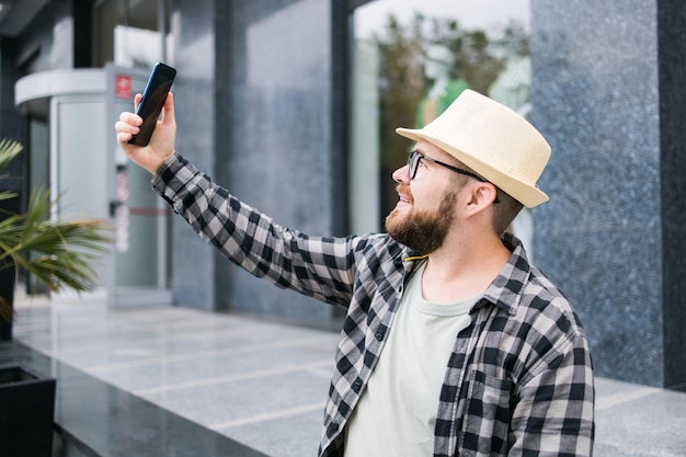 Bebaarde man neemt selfie via smartphone wandelingen in het centrum poses tegen moderne stad gebouw geniet van vrije tijd mensen stedelijke levensstijl en technologie concept