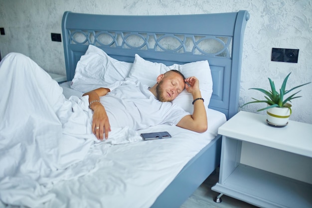 Bebaarde man slaapt alleen op een groot en gezellig bed wit beddengoed met smartphone