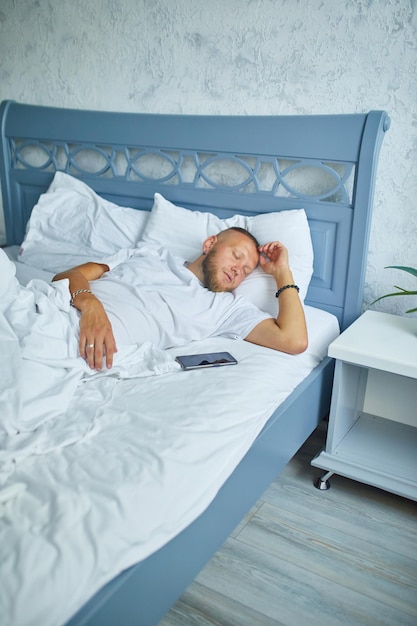 Bebaarde man slaapt alleen op een groot en gezellig bed wit beddengoed met smartphone