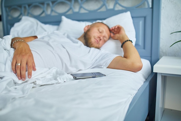 Bebaarde man slaapt alleen op een groot en gezellig bed wit beddengoed met smartphone