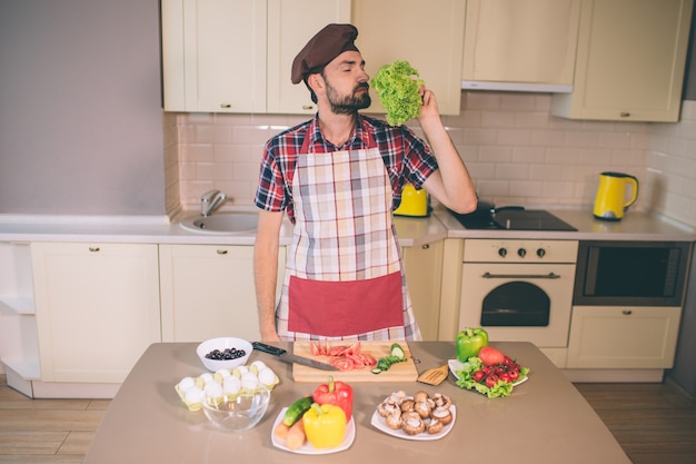 Bebaarde man staat en ruikt bladeren van letuce. Hij houdt de ogen gesloten. De mens is serieus en geconcentreerd. Guy staat in de keuken. Hij bereidt eten.