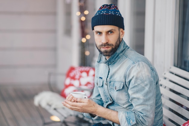 Bebaarde man van middelbare leeftijd met blauwe ogen dikke baard en snor draagt een stijlvolle hoed en een spijkerjasje houdt een kop hete thee of koffie vast heeft een doordachte uitdrukking denkt aan iets belangrijks