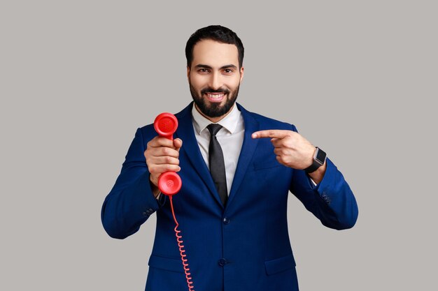 Bebaarde man wijzende vinger op handset van rode vintage vaste telefoon geïnteresseerd in retro apparaten beantwoorden van oproepen dragen officiële stijl pak Indoor studio shot geïsoleerd op grijze achtergrond