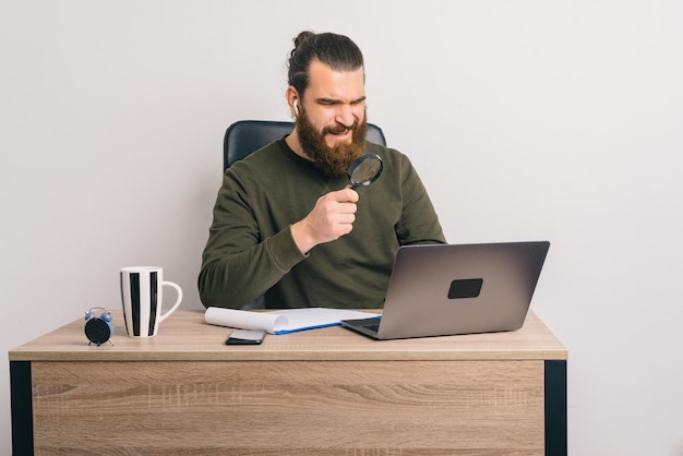 Bebaarde man zit aan het bureau en kijkt door een vergrootglas naar de laptop.