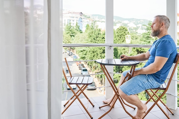 Bebaarde man zittend aan houten tafel op hotelbalkon geniet van zomervakantie