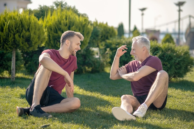 Bebaarde mannelijke en grijsharige man zittend op het gras, ontspannen na de training