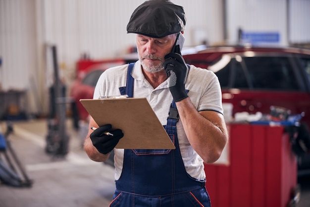 Bebaarde mannelijke werknemer die aantekeningen controleert terwijl hij een telefoongesprek voert bij het autoreparatieservicestation