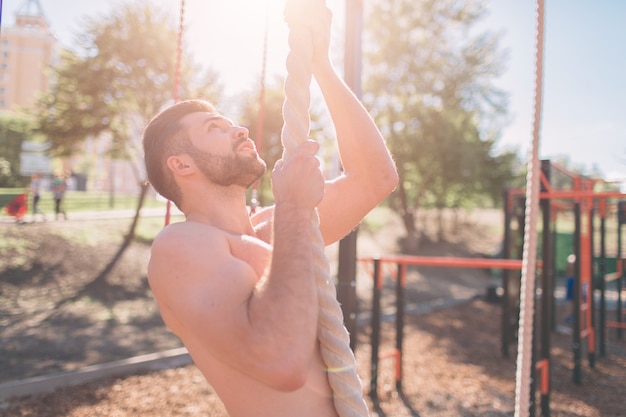 Bebaarde met zwart haar man dik touw klimmen tijdens fitness oefeningen in cross-fit training met