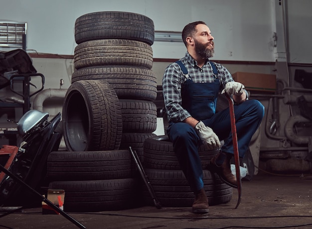 Bebaarde monteur gekleed in een uniform, houdt een koevoet vast, zit op oude autobanden in de garage.