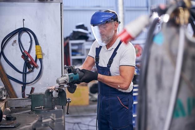 Bebaarde monteur in beschermend masker met grinder in garage