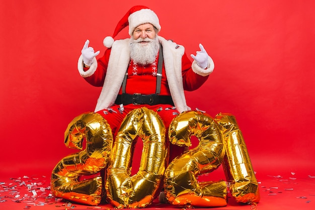 bebaarde oude man in Santa Claus-kostuum dichtbij gouden baloons van 2021 die op rode achtergrond worden geïsoleerd