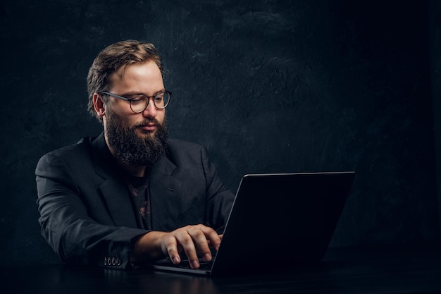 Bebaarde programmeur in pak werkt op laptop zittend aan tafel op kantoor tegen een donkere muur