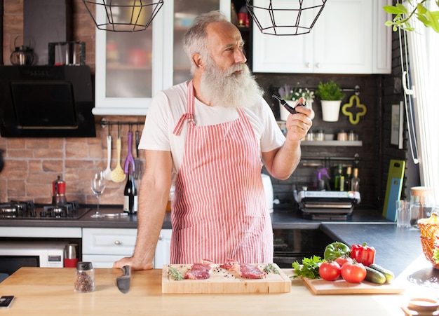 Bebaarde senior man koken steaks op zijn keuken.