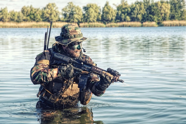 Bebaarde soldaat van speciale troepen in actie tijdens rivieroverval in het jungle-terrein. Hij is tot zijn middel diep in het water en de modder en klaar om de vijand te ontmoeten, te overleven en te vechten in een agressieve vijandige omgeving