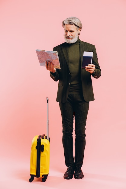 Bebaarde stijlvolle man van middelbare leeftijd met een gele koffer, kaart en paspoort met een kaartje op roze.