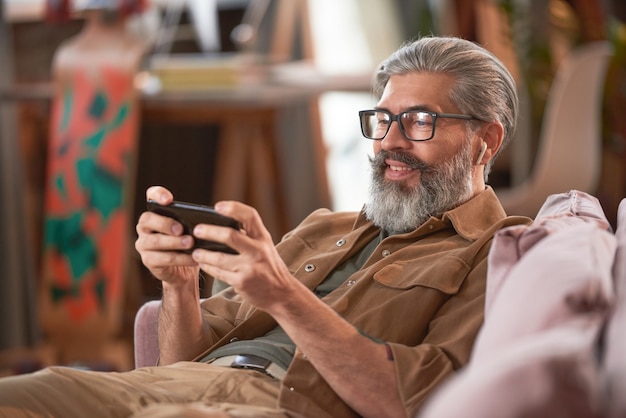 Bebaarde volwassen man in bril die online games speelt op zijn mobiele telefoon terwijl hij op de bank in de kamer zit