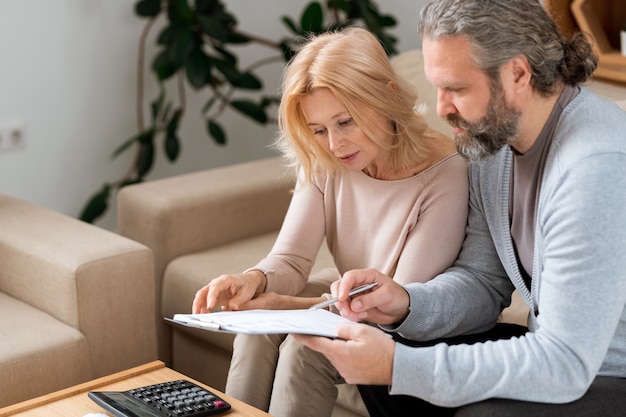 Bebaarde volwassen man wijst op contract voor het kopen van een nieuw huis terwijl hij het leest met zijn vrouw voordat hij ondertekent