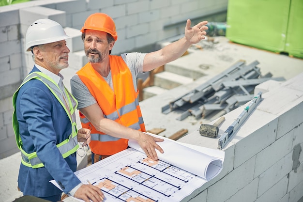 Bebaarde volwassen mannelijke werknemer in een veiligheidshelm wijzend op iets naar een tevreden ingenieur